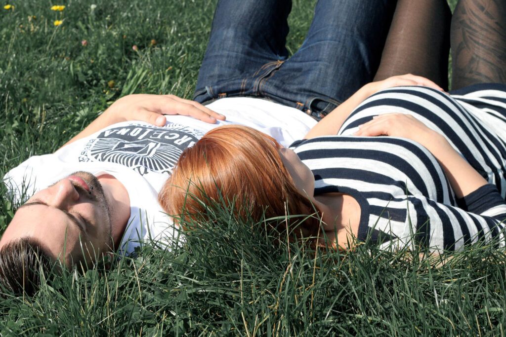 Ein glückliches schwangeres Paar liegt im grünen Rasen und genießt die Wärme der Sonne und die Zweisamkeit während ihres Schwangerschaftsshootings im Freien.