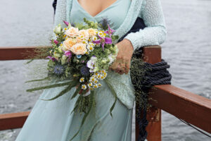 Romantische Umarmung: Der Bräutigam umarmt die Braut von hinten, während sie ihre Hände verschränken. Der Brautstrauß steht im Fokus, und das Bild wurde auf einem Floß aufgenommen.
