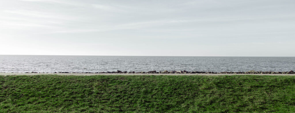 Symmetrisches Foto von der Ostsee die horizontal von Links nach rechts verläuft. Zu sehen ist der asphaltierte Fuß- und Radweg und der grüne Deich. #faq #fotografie #datenschutz