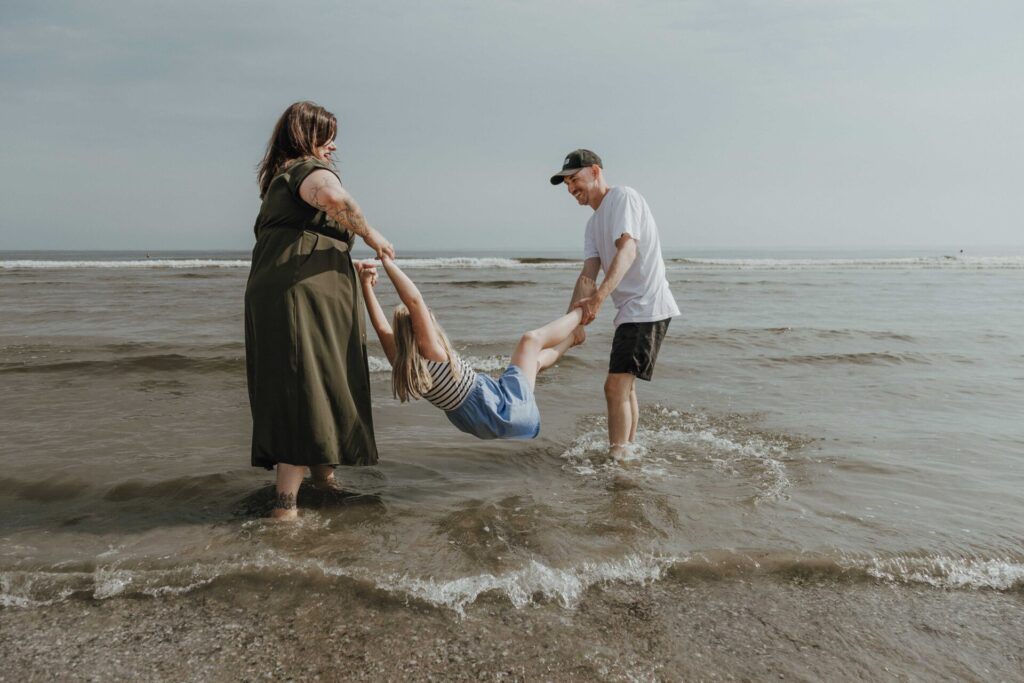 Ein blondes Mädchen hängt fröhlich zwischen ihren Eltern in der Ostsee – die Mutter hält ihre Arme, der Vater ihre Füße, und alle lachen ausgelassen.