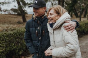 Ein Paar in Winterjacken spaziert lachend die Promenade in Binz entlang. Sie schließt die Augen, während er sie liebevoll im Arm hält.