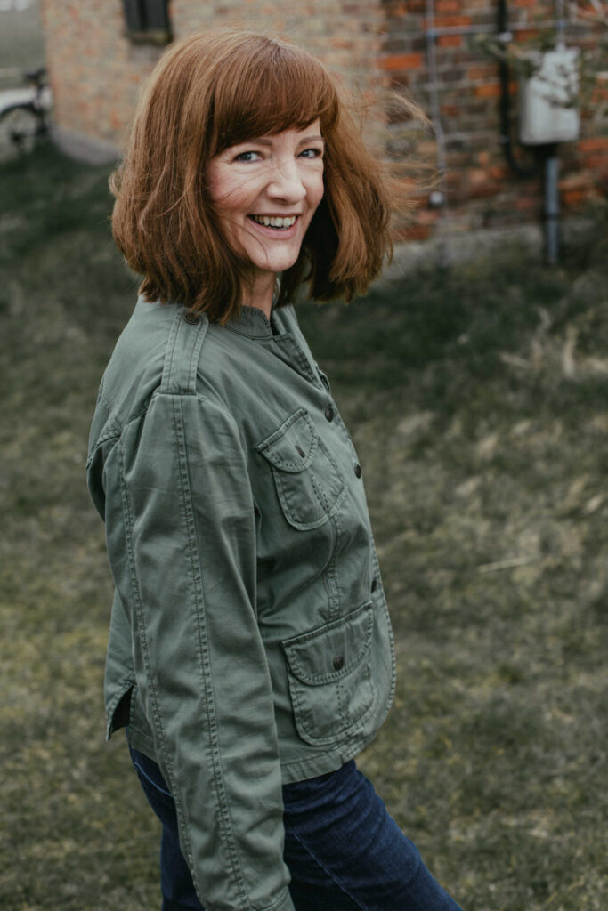 Eine Frau mit kupferroten Haaren trägt eine mintgrüne Jacke und lacht in die Kamera. Im Hintergrund ist ein Backsteinhaus und grüner Rasen zu sehen.