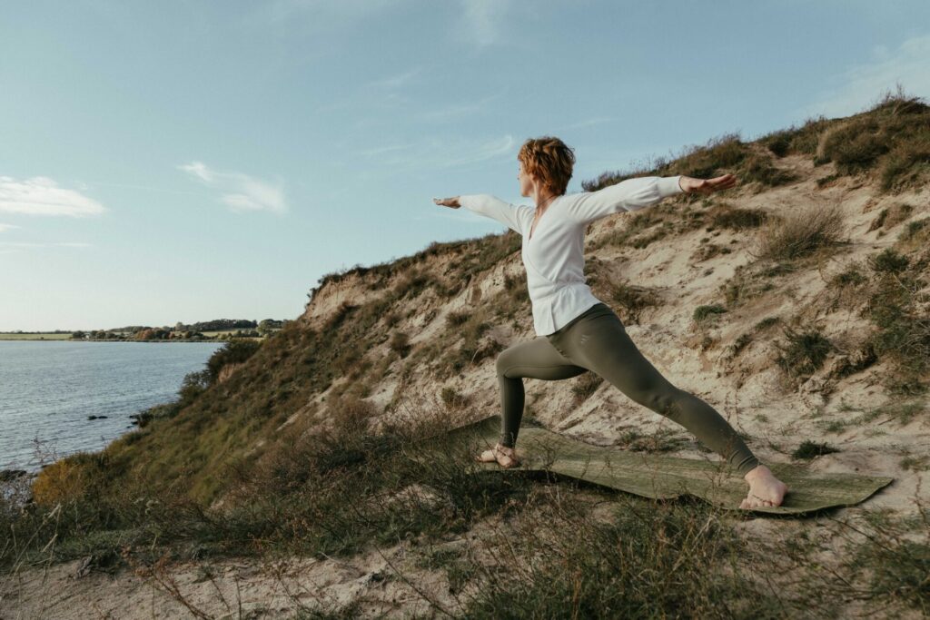 Eine Frau steht bei dieser Portraitfotografie auf Rügen in der Yogapose der Krieger auf einem sandigen Hügel mit Blick in die Ferne auf das Meer.