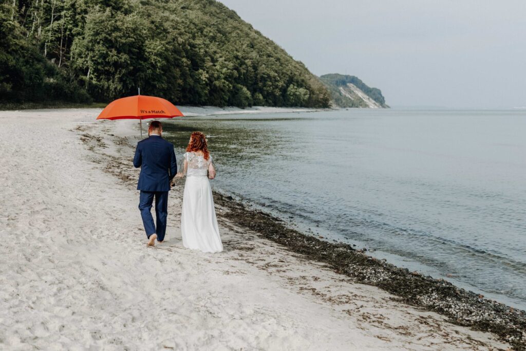 Hochzeitspaar am Strand auf Rügen mit rotem Regenschirm. Hochzeitsfotografie in Sellin auf Rügen von Franziska Lorenz.