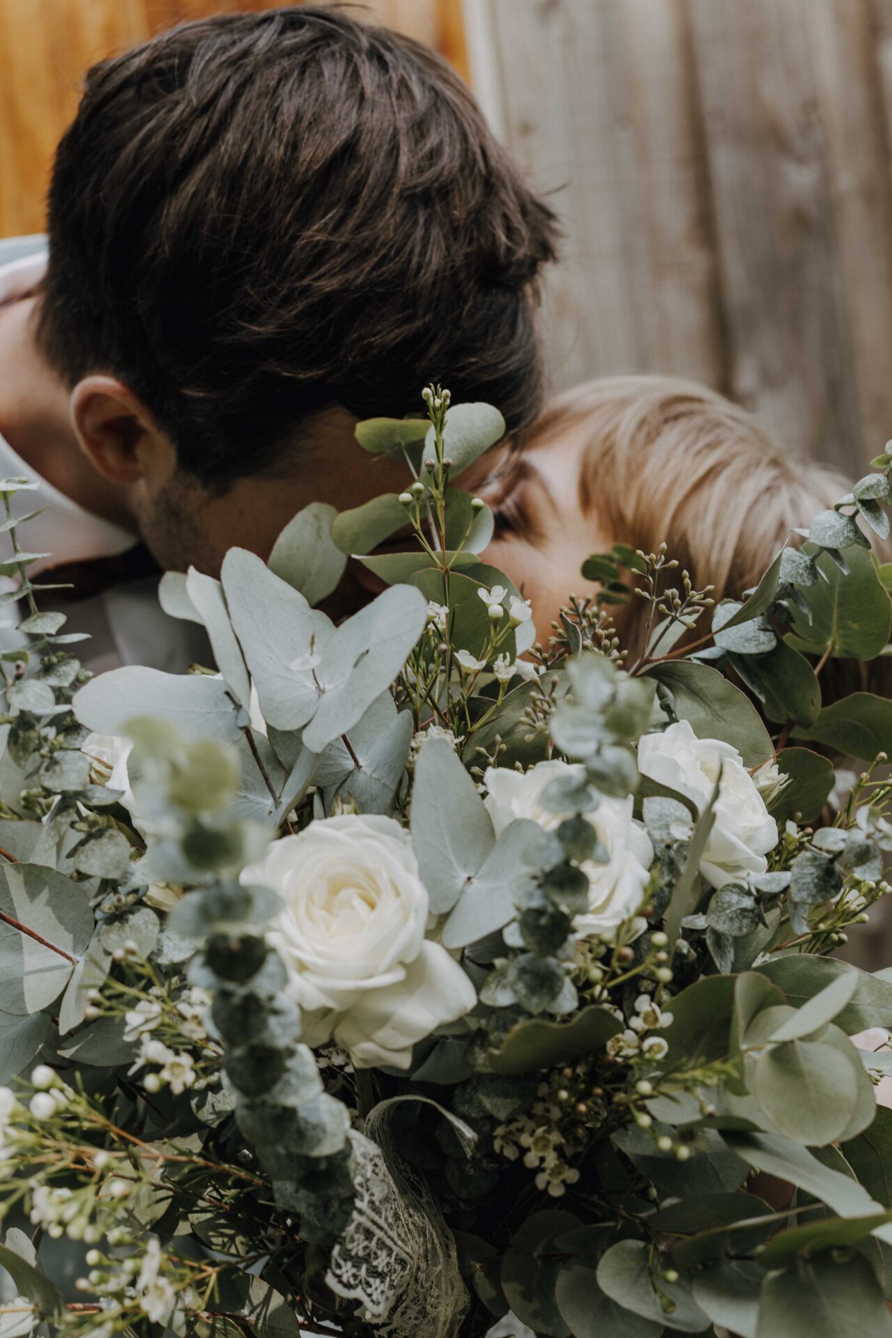 Foto des küssenden Brautpaares, das sich hinter einem prachtvollen Brautstrauß aus Eukalyptus und weißen Rosen versteckt.
