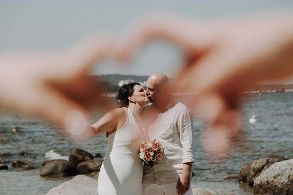 Kuss eines Brautpaares am Strand in Binz auf Rügen, eingerahmt durch ein Herz aus Händen – Hochzeitsfotografie von Franziska Lorenz