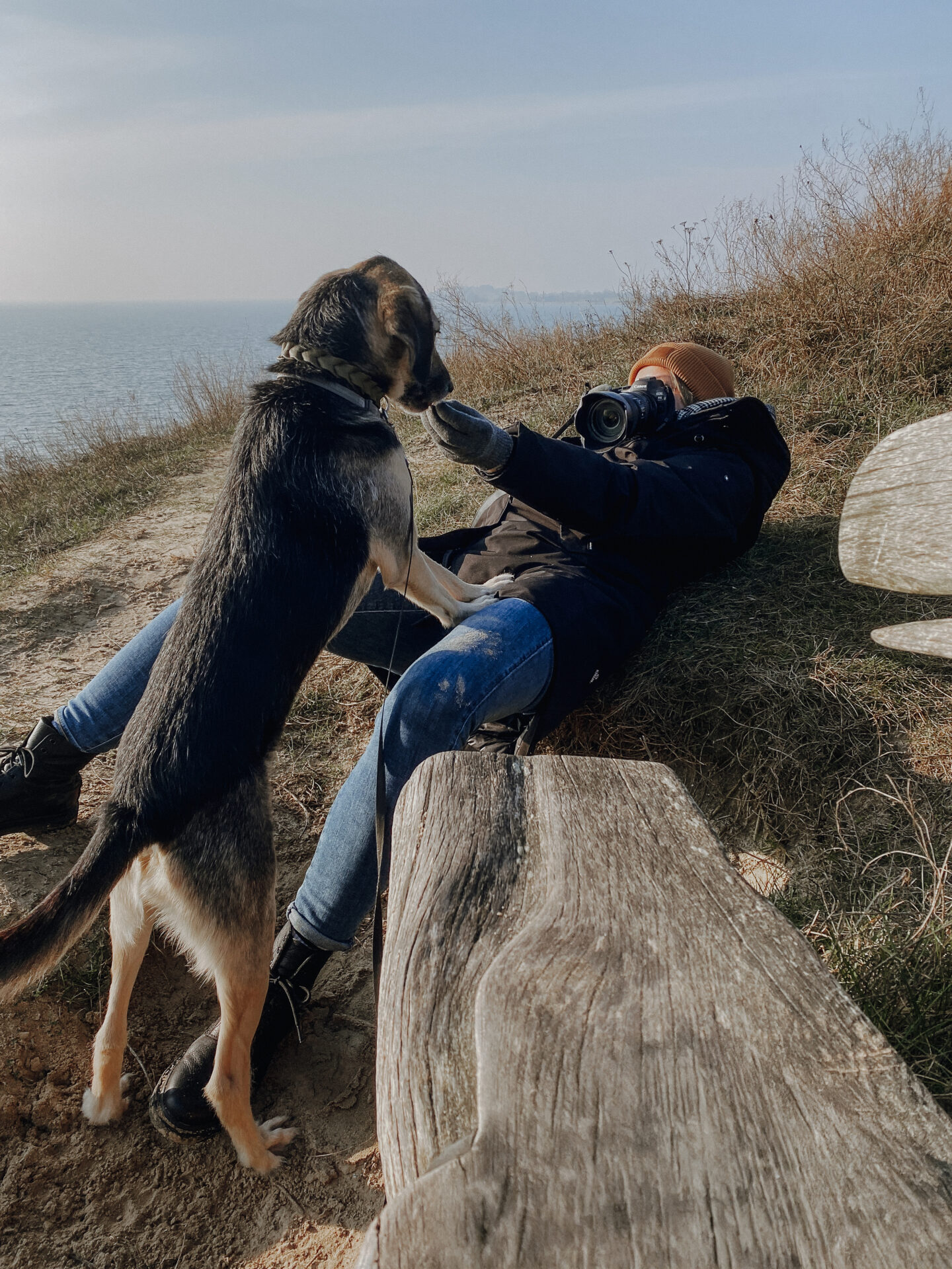 Dynamische Haustierfotografie, Hundefotografie, Tierfotografie auf Rügen: Fotografin liegt auf dem Boden, ein mittelgroßer Misschlingshund und hat die Pfoten auf ihrem Knie abgestützt und bekommt ein Leckerli von ihr. Im Hintergrund sieht man den Horizont der Hagenschen Wiek in Mönchgut auf Rügen